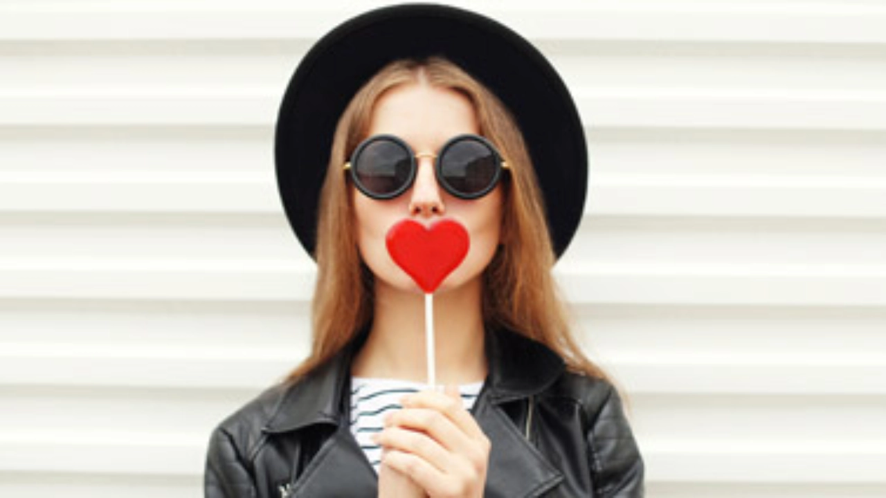Junge Frau mit Sonnenbrille hält sich einen Lolly in Herzform vor den Mund.
