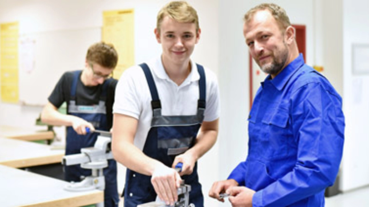 Azubi steht mit seinem Ausbilder an einem Schraubstock und feilt etwas.