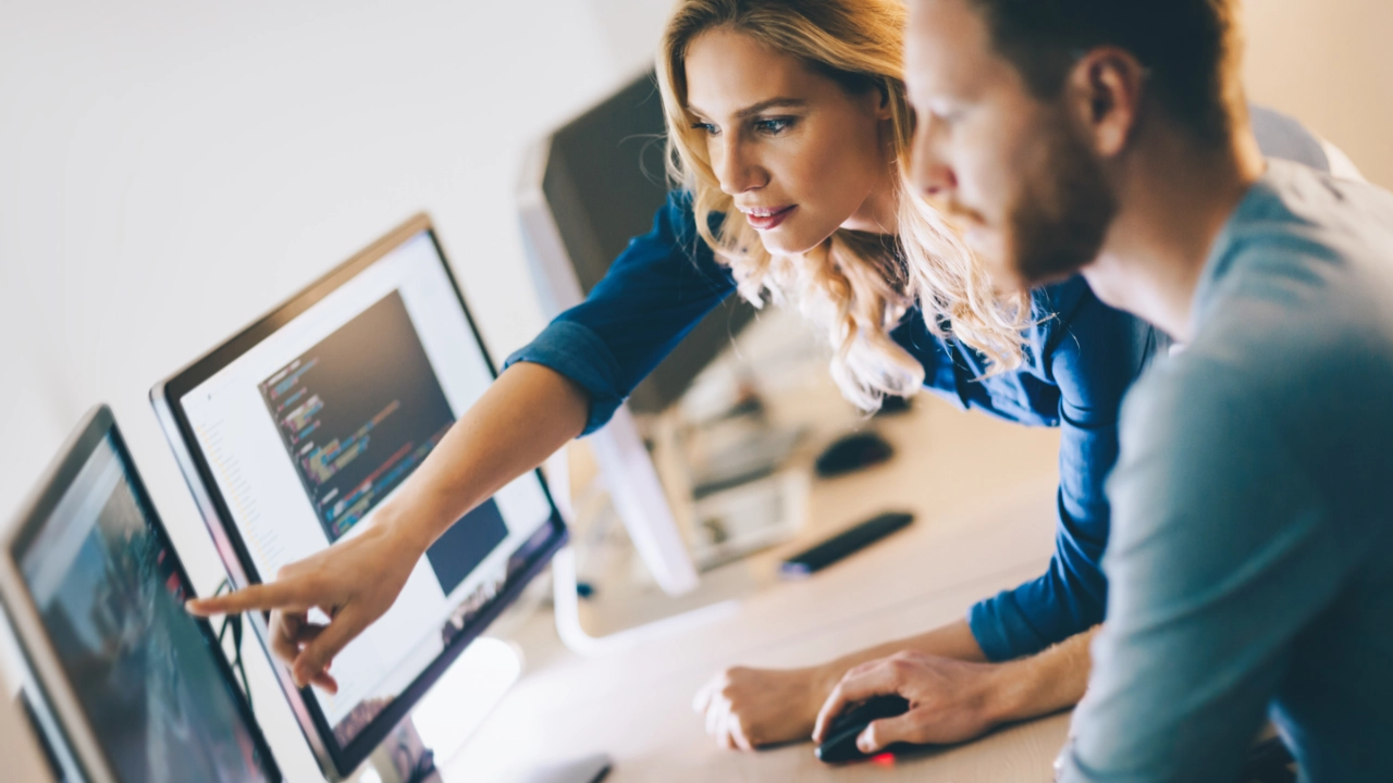 Eine Frau zeigt mit ihrem Finger auf einen PC Monitor und ein Mann Vordergrund schaut in die gezeigte Richtung 