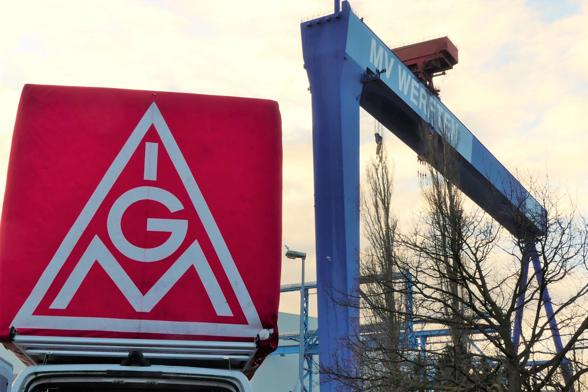 Im Vordergrund steht ein Tranporter mit einem großen IG Metall Würfel drauf und auf der rechten Bildhälfte ist ein großer Brückenkran mt dem MV-Werften Schriftzug