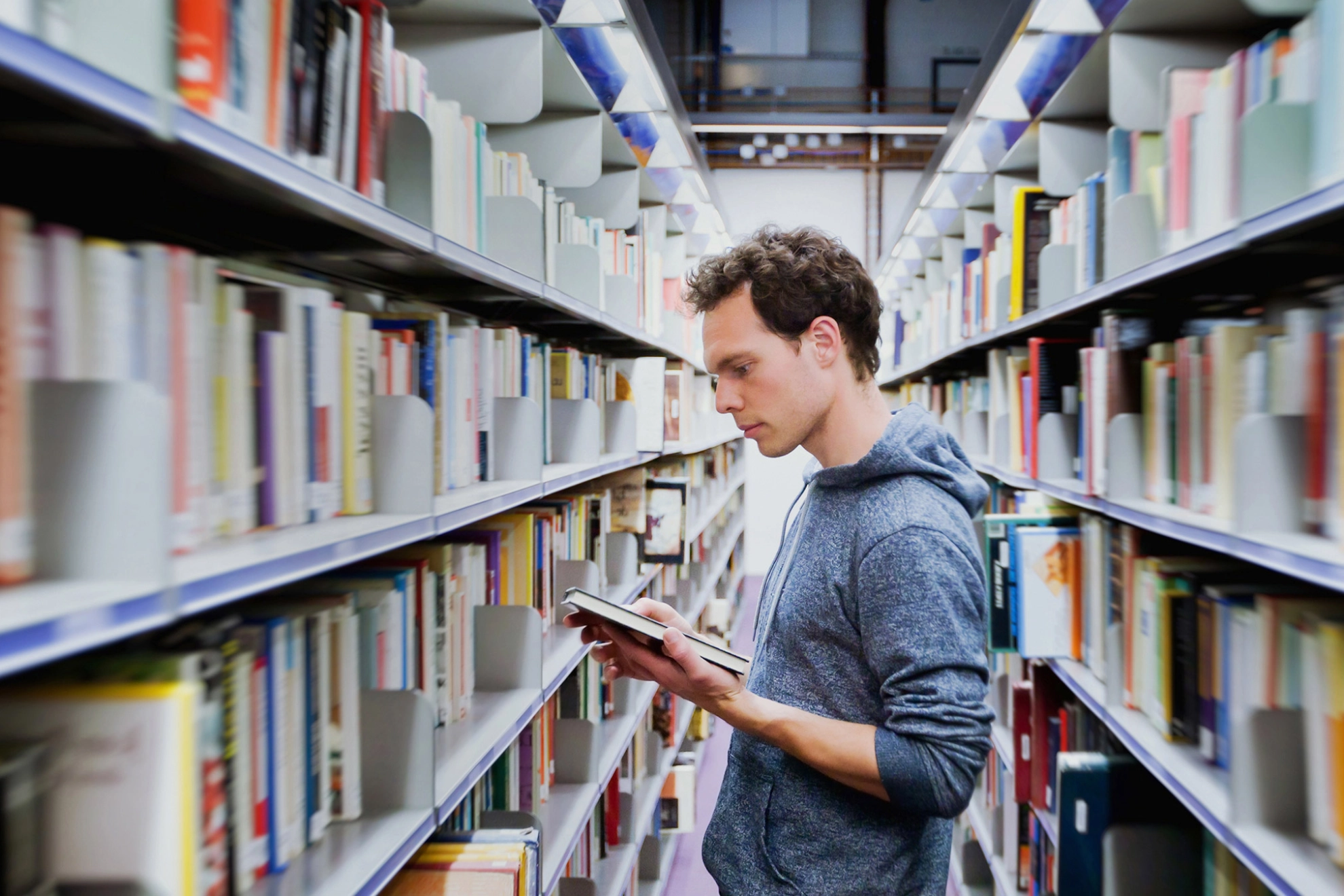Junger Mann steht zwischen Regalen in einer Bibliothek und schaut in ein Buch.