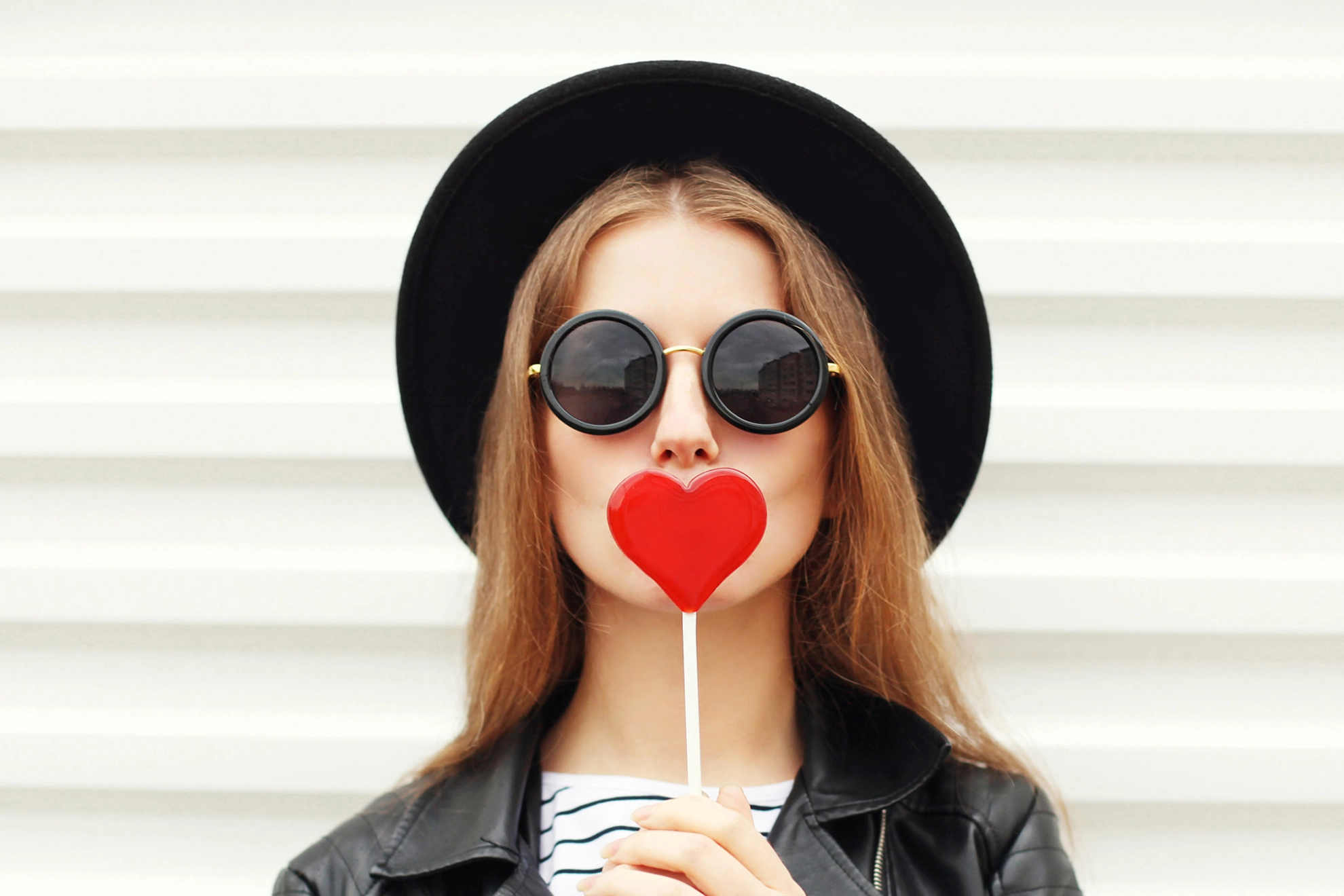 Junge Frau mit Sonnenbrille hält sich einen Lolly in Herzform vor den Mund.
