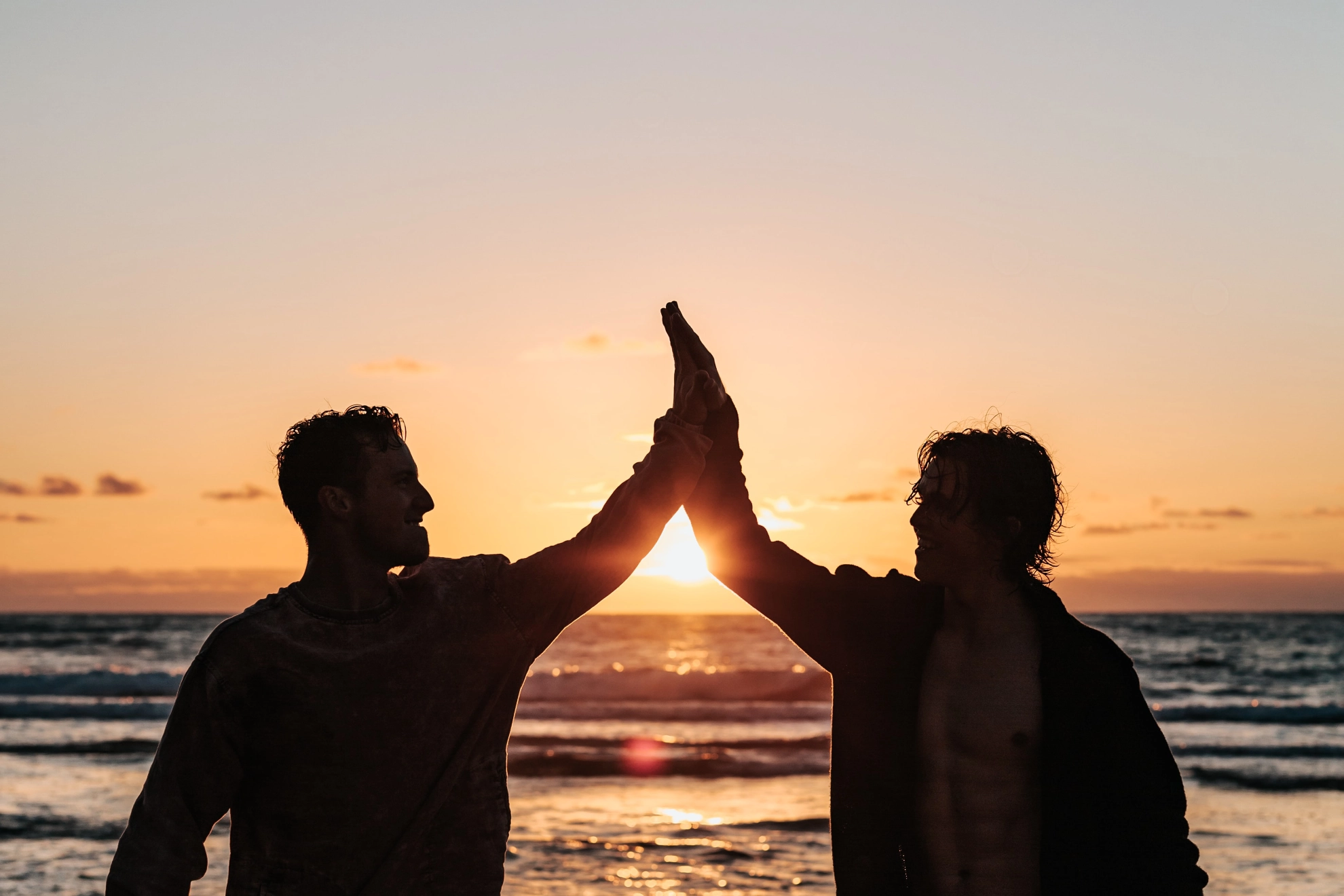 Zwei Menschen geben sich ein Highfive, am Strand bei Sonnenuntergang.