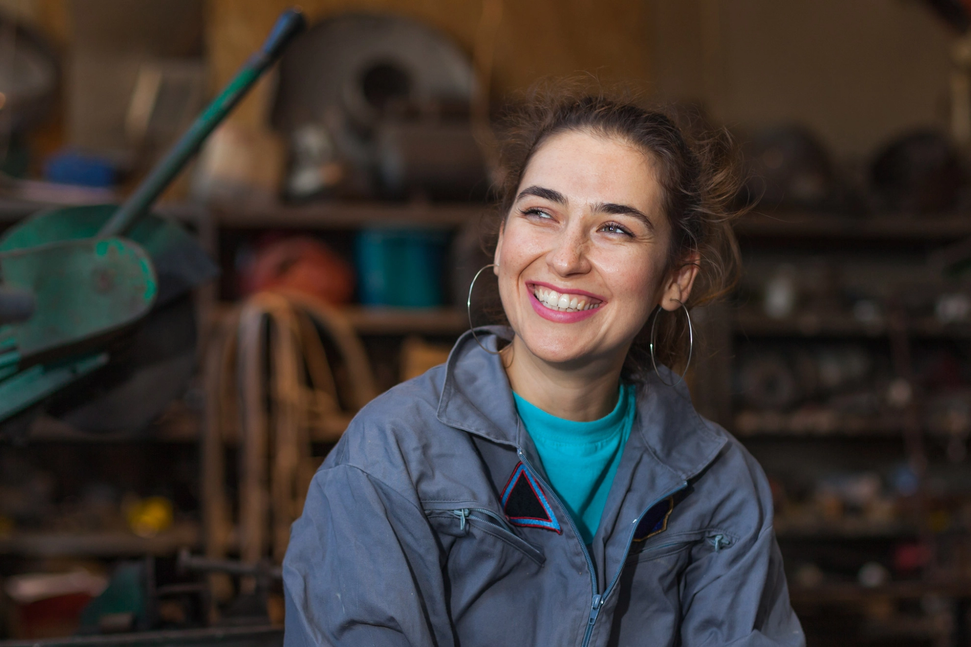 Junge Frau im Blaumann sitzt lächelnd in einer Werkstatt.