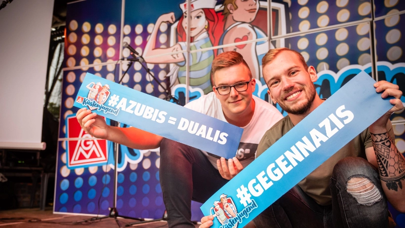 Zwei Jungs halten je eine Tafel mit den Hashtags: "gegen Nazis" und "Azubis = Dualis"