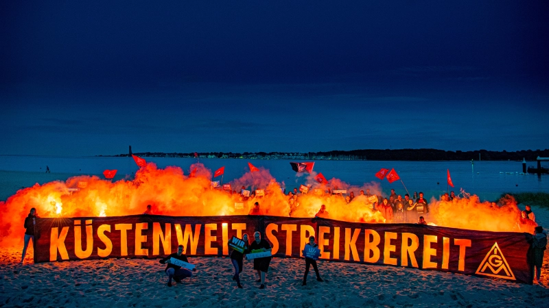 Ein Gruppe junge Leute halten ein sehr langes Transarent mit der Aufschrift: "Küstenweit Streikbereit" bei einer abdendlichen Stimmung. Hinten den Menschen fackeln Bengalische rote Lichter ab, die wirken als wenn es Flammen wären.